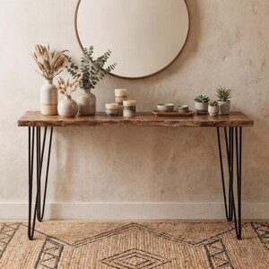 Op de afbeelding: Een rechthoekige houten sidetable met een natuurlijke boomstamrand en vier zwarte metalen haarspeldpoten. Het tafelblad bevat drie keramische vazen met gedroogde takken, drie stompkaarsen, een houten dienblad met schaaltjes en drie vetplantjes.