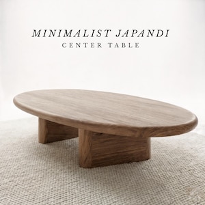 May include: A wooden centre table with an oval top and two rectangular supports. The table is a natural wood colour and sits on a light-coloured rug. The text "MINIMALIST JAPANDI CENTER TABLE" is at the top.