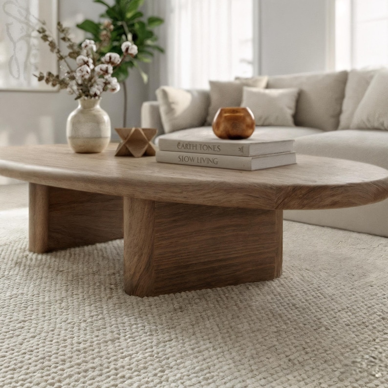 May include: An oval wooden coffee table in a light brown hue. The table features a solid base and is styled with books, a decorative object, a vase with cotton stems, and a copper-coloured candle holder. The books read "EARTH TONES" and "SLOW LIVING".