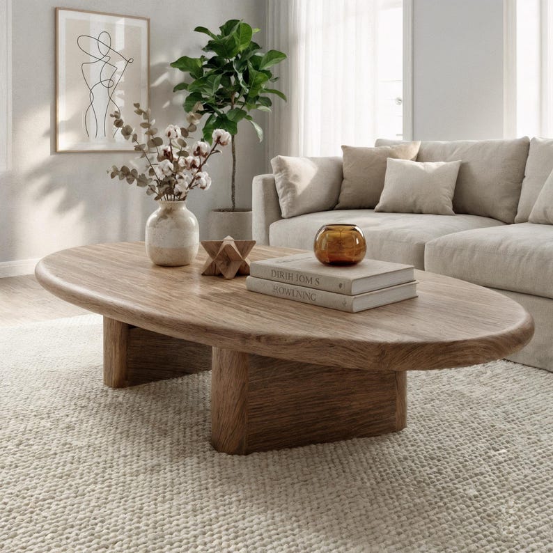 May include: An oval wooden coffee table in light brown, with a natural finish. The table is supported by two rectangular legs. Books and a glass bowl are on top. The room includes a beige sofa, a rug, and a framed art print.