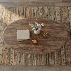 May include: An oval wooden coffee table with a natural wood grain. It features a small white vase with greenery and white flowers, a stack of beige napkins, a gold star, and a small candle in a glass holder. The table sits on a patterned rug.