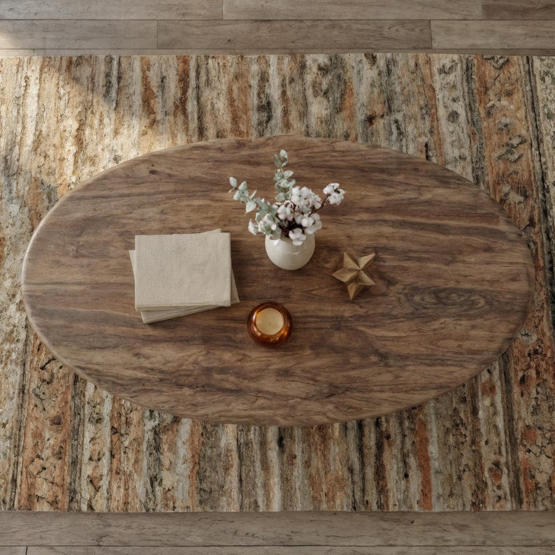 May include: An oval wooden coffee table with a natural wood grain. It features a small white vase with greenery and white flowers, a stack of beige napkins, a gold star, and a small candle in a glass holder. The table sits on a patterned rug.