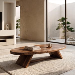 May include: An oval coffee table crafted from solid brown wood with a natural grain pattern, featuring a thick tabletop and two angled support legs. Two stacked books and a small ceramic mug sit on the surface of the wooden table.