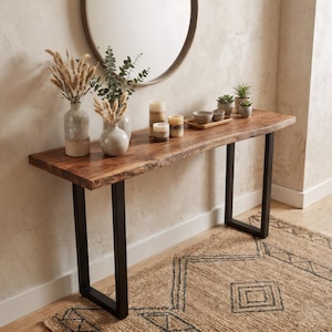 Puede incluir: Una consola de madera con una tapa de borde vivo y patas de metal negro. La mesa está decorada con jarrones, velas y pequeñas plantas en macetas. Un espejo redondo cuelga de la pared encima de la mesa y una alfombra estampada está en el suelo.