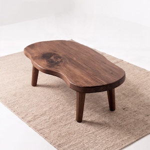 Op de afbeelding: Een lage salontafel van donkerbruin hout met een natuurlijke, onregelmatige niervorm. Het tafelblad heeft een opvallende houtnerf en een zichtbare knoest, ondersteund door drie stevige, cilindervormige houten poten.