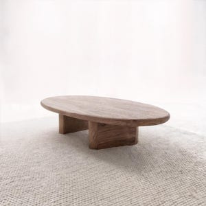 Puede incluir: Una mesa de centro ovalada de madera con un acabado natural. La tapa de la mesa está soportada por dos patas rectangulares. La mesa está sobre una alfombra clara y texturizada, creando una estética minimalista.