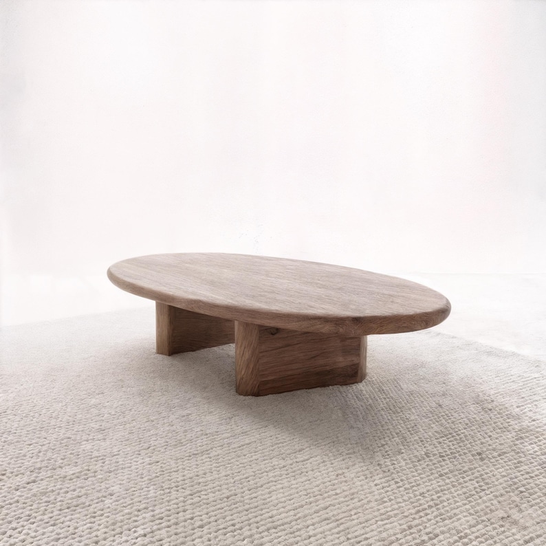 May include: A low, oval wooden coffee table with a natural finish. The tabletop is supported by two rectangular legs. The table sits on a light-coloured, textured rug, creating a minimalist aesthetic.
