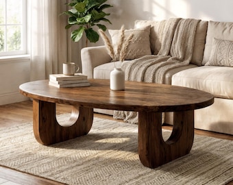 May include: An oval coffee table crafted from dark brown wood with a smooth surface and two U-shaped pedestal legs. The table is positioned on a textured beige rug and holds a small white ceramic vase containing dried grass and a stack of books.