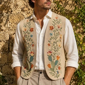 Puede incluir: Un chaleco beige sin mangas con intrincados bordados de cactus verdes y flores coloridas en tonos naranja, rojo y azul en los paneles frontales, usado sobre una camisa blanca de manga larga con botones y pantalones de color claro.