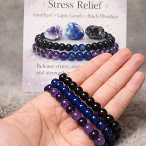 May include: Three beaded bracelets in purple, blue, and black are shown. The bracelets are made of smooth, round beads. A card behind the bracelets reads "Stress Relief" and lists the stones: Amethyst, Lapis Lazuli, and Black Obsidian.