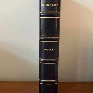 May include: A dark blue book titled "Naval Gunnery" by Douglas, with gold lettering. The spine has gold horizontal lines and the year 1829 at the bottom. The book is on a wooden surface.