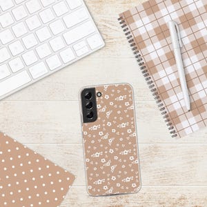 May include: A clear phone case with a floral pattern of small white flowers on a tan background. The case is lying on a light wood-grain surface, next to a white keyboard, a notebook, and a pen.