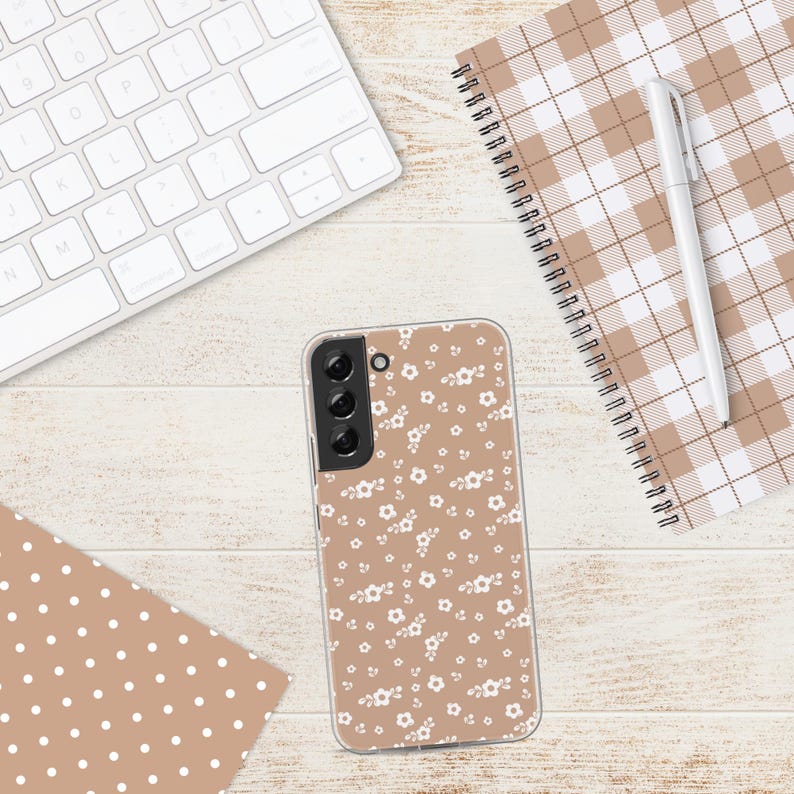 May include: A clear phone case with a floral pattern of small white flowers on a tan background. The case is lying on a light wood-grain surface, next to a white keyboard, a notebook, and a pen.