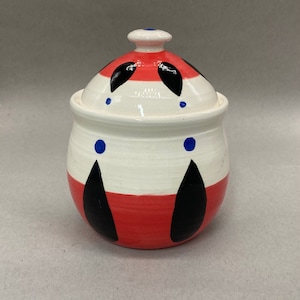 Puede incluir: Un azucarero de cerámica blanca con un diseño geométrico rojo y negro. La tapa tiene un pomo en la parte superior y un diseño rojo y negro.