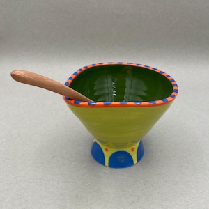 Puede incluir: Un cuenco de cerámica pintado a mano con una cuchara de madera. El cuenco es de color verde lima con un interior verde y un borde azul y naranja. La base es azul y amarilla. La cuchara es marrón claro.