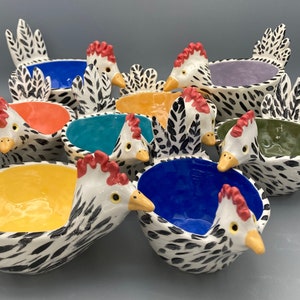 May include: A collection of ceramic chicken-shaped bowls with various interior colors, including blue, orange, yellow, and green. The chickens have white bodies with black spots, red combs, and yellow beaks. The bowls are arranged on a gray surface.