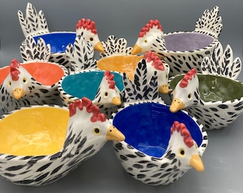 Puede incluir: Una colección de cuencos de cerámica con forma de gallina, cada uno con un cuerpo blanco con patrones de pinceladas negras, una cresta roja y un pico amarillo. El interior de cada cuenco está esmaltado en un color sólido diferente, incluyendo azul, naranja, amarillo y verde.