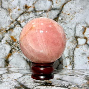 May include: A large, polished rose quartz sphere, displaying a soft pink hue with natural veining. The sphere rests on a dark brown, wooden, decorative stand. The background features a marble-like pattern.