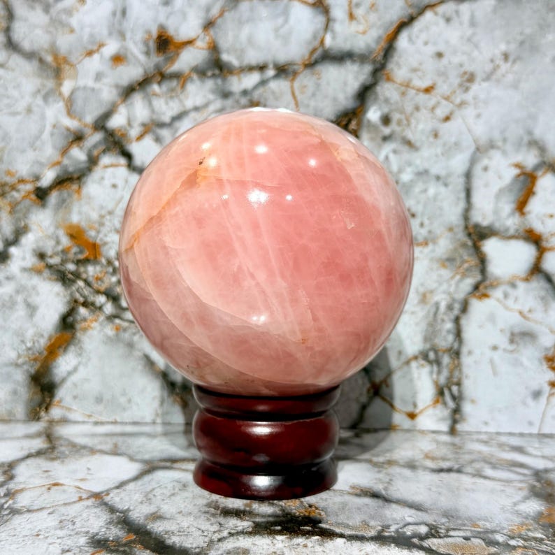 May include: A large, polished rose quartz sphere, displaying a soft pink hue with natural veining. The sphere rests on a dark brown, wooden, decorative stand. The background features a marble-like pattern.