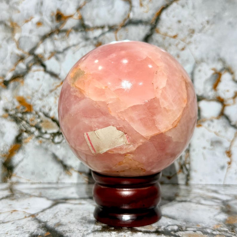 May include: A large, polished sphere of pink rose quartz, resting on a dark wooden stand. The sphere has a mottled appearance with varying shades of pink and some areas of lighter color. A small, partially visible label is affixed to the sphere.