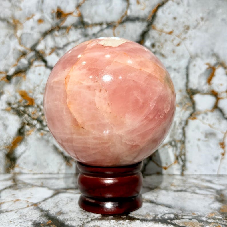 May include: A large, polished rose quartz sphere, displaying a soft pink hue with subtle variations in tone. The sphere rests on a dark, varnished wooden stand. The stone's surface reflects light, showcasing its natural patterns.