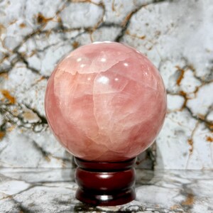 May include: A large, polished rose quartz sphere, displaying a soft pink hue with natural veining. The sphere rests on a dark brown, wooden stand, creating a striking contrast. This decorative crystal ball is a unique home decor item.