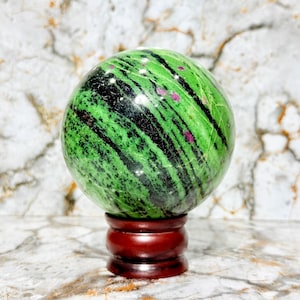 Puede incluir: Una esfera pulida de zoisita verde con vetas negras y pequeñas inclusiones de rubí rojo. La bola de piedra preciosa se exhibe sobre un soporte de madera rojo oscuro, sobre un fondo de mármol. Un artículo de decoración único.