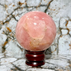 May include: A large, polished rose quartz sphere, displaying a soft pink hue with lighter, translucent patches. The sphere rests on a dark, reddish-brown wooden stand. The background features a marble-like pattern.