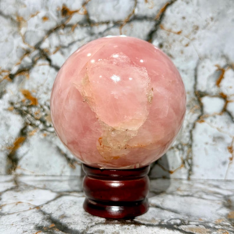 May include: A large, polished rose quartz sphere, displaying a soft pink hue with lighter, translucent patches. The sphere rests on a dark, reddish-brown wooden stand. The background features a marble-like pattern.
