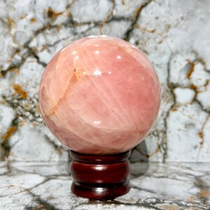 May include: A large, polished rose quartz sphere, displaying a soft pink hue with subtle variations in tone. The sphere rests on a dark, reddish-brown wooden stand, set against a backdrop of a marble-patterned surface. This decorative item is ideal for home decor.