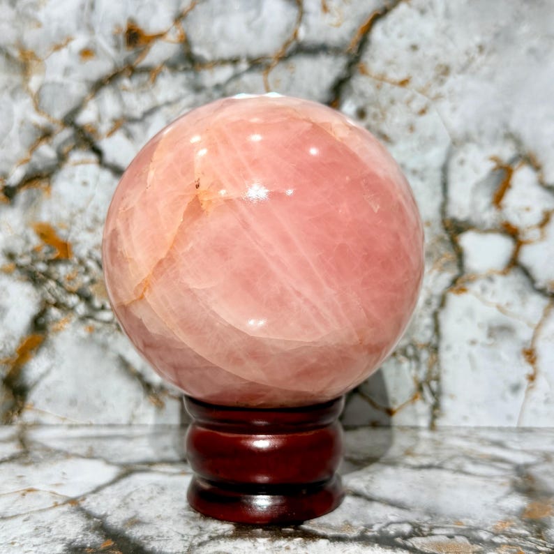 May include: A large, polished rose quartz sphere, displaying a soft pink hue with subtle variations in tone. The sphere rests on a dark, reddish-brown wooden stand, set against a backdrop of a marble-patterned surface. This decorative item is ideal for home decor.