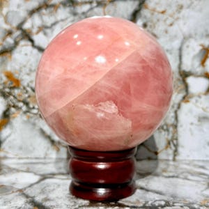 May include: A large, polished rose quartz sphere, displaying a soft pink hue with natural veining. The sphere is mounted on a dark, reddish-brown wooden stand, creating a striking contrast. This decorative item is ideal for home decor.