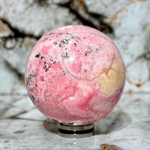May include: A polished sphere of rhodochrosite, showcasing a blend of pink hues with black and beige inclusions. The gemstone ball is mounted on a silver-colored base, creating a striking contrast. The sphere is a decorative item.