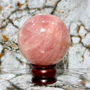 May include: A large, polished rose quartz sphere, displaying a soft pink hue with natural veining. The sphere rests on a dark brown, wooden stand. The background features a marble-like pattern.
