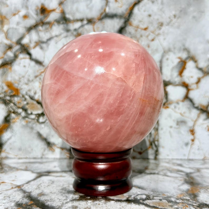 May include: A large, polished rose quartz sphere, displaying a soft pink hue with natural veining. The sphere rests on a dark brown, wooden stand. The background features a marble-like pattern.