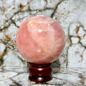 May include: A large, polished rose quartz sphere, displaying varying shades of pink and a translucent quality. The crystal ball sits atop a dark, reddish-brown wooden stand, with a marble-patterned background.