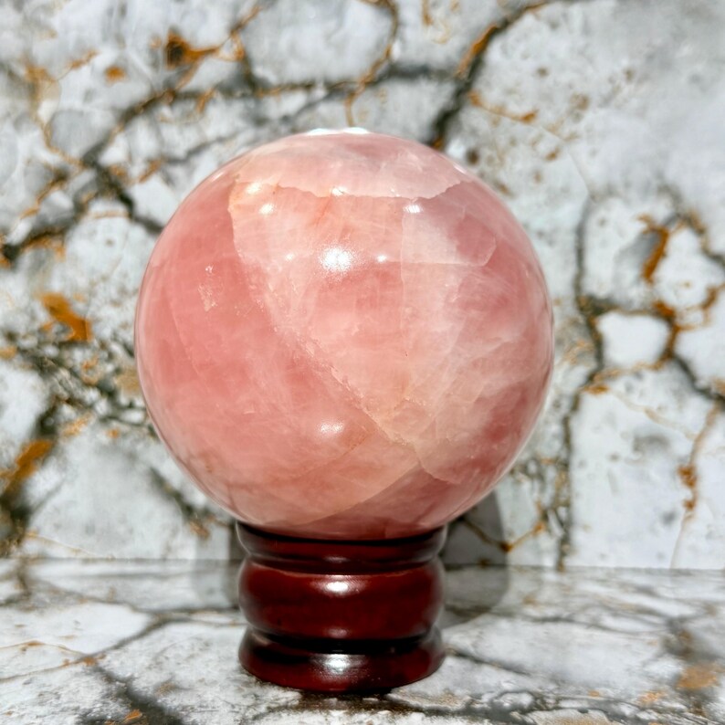 May include: A large, polished rose quartz sphere, displaying varying shades of pink and a translucent quality. The crystal ball sits atop a dark, reddish-brown wooden stand, with a marble-patterned background.