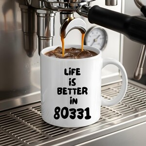 May include: A white ceramic mug with a handle, featuring the black printed text "LIFE IS BETTER IN 80331" on the front. The mug is filled with a dark brown liquid and ice cubes, positioned directly under the spout of a stainless steel espresso machine.