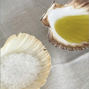 May include: Two seashell-shaped dishes on a grey linen surface. One shell holds coarse white salt, the other contains yellow liquid. The shells have a cream and brown colour scheme, suggesting a coastal or nautical theme.