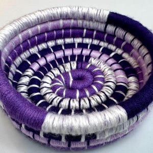 May include: A small, round, coiled basket crafted from yarn in shades of deep purple, lavender, and white. The structure features a continuous spiral pattern held together by vertical stitching, creating a textured, bowl-shaped container.