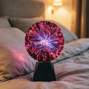 Może przedstawiać: Sferyczna lampa plazmowa z czarną podstawą i szklaną kulą wypełnioną świecącymi różowymi i fioletowymi włóknami elektrycznymi, które promieniują ze środka, tworząc dynamiczny pokaz świetlny wewnątrz przezroczystej, okrągłej szklanej struktury.