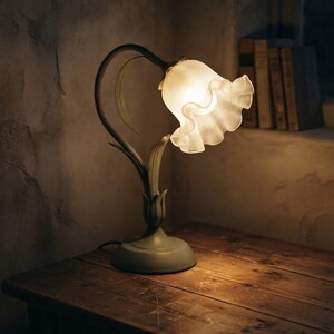 May include: A table lamp with a green metal base shaped like a flower stem and leaves. The lamp features a single, downward-facing, ruffled, frosted glass shade that emits a warm, soft glow, resembling the appearance of a blooming flower.