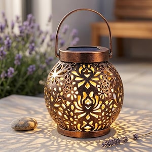 Peut inclure: Une lanterne solaire en bronze avec un motif floral ajouré. La lanterne est dotée d'une poignée et est posée sur une surface en pierre, projetant des ombres décoratives. Une petite pierre et une brindille de lavande sont également visibles.