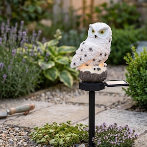 May include: A white owl garden stake with black spots and yellow eyes. The owl is perched on a brown base with a solar panel attached. The stake is black and is placed in a garden setting with plants and flowers.