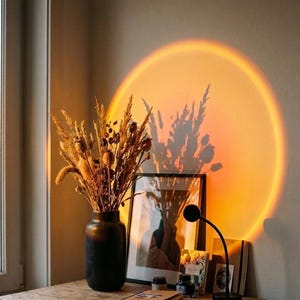 Peut inclure: Un vase noir rempli d'herbes séchées et de gousses de graines est placé à côté d'une impression encadrée et d'une petite lampe noire qui projette une grande lueur circulaire orange chaud rappelant un coucher de soleil sur le mur.