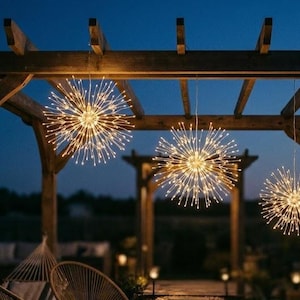 May include: Three spherical starburst lights with warm white LED filaments suspended from a wooden pergola. Each light features a central core with numerous thin, radiating wire branches that create a glowing, firework-like effect at dusk.
