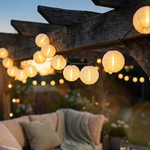 Op de afbeelding: Een lichtsnoer met ronde, witte papieren lantaarns die een warm, zacht schijnsel verspreiden, opgehangen aan een donkere houten pergola. De bolvormige lampjes zijn gelijkmatig verdeeld over een dunne zwarte draad voor verlichting.