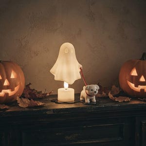 Op de afbeelding: Een witte spookvormige lamp met een oplichtende voet, verbonden door een rode riem aan een klein wit puppybeeldje. Het spook en de puppy staan op een zwarte basis, geflankeerd door twee uitgesneden oranje pompoenen en verspreide bruine bladeren.