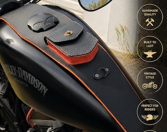 May include: A black leather motorcycle tank bib with orange trim and an attached small storage pouch featuring a brass twist lock. The bib is mounted on a black fuel tank with a skull-shaped gas cap and includes a small metal D-ring attachment.