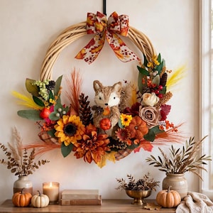 May include: A circular wreath made of light brown woven branches, decorated with a central plush fox figure, orange sunflowers, dark orange dahlias, beige roses, pinecones, green leaves, and a patterned bow with autumn leaves at the top.